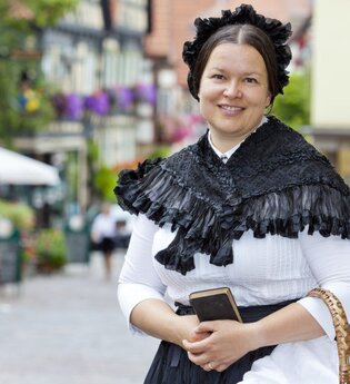 Frau in historischer Kleidung mit schwarzem Umhang und Haube in einer malerischen Altstadt. | © Stadt Besigheim