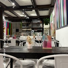 Ein stilvoll eingerichtetes Café mit modernen Kunstwerken an den Wänden und weißen Stühlen um dunkle Holztische. | © see on the left