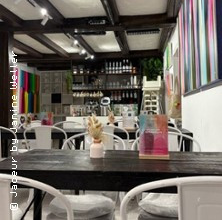 Ein stilvoll eingerichtetes Café mit modernen Kunstwerken an den Wänden und weißen Stühlen um dunkle Holztische. | © see on the left