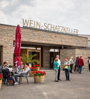 Einladende Szene vor einem Gebäude. Gäste sitzen entspannt an Tischen, umgeben von blühenden Blumenkübeln. Eine Gruppe steht am Eingang. Die Fassade ist aus Naturstein und auf dem Dach steht die Aufschrift "Wein-Schatzkeller" in goldenen Großbuchstaben.