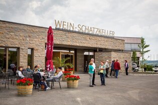 Einladende Szene vor einem Gebäude. Gäste sitzen entspannt an Tischen, umgeben von blühenden Blumenkübeln. Eine Gruppe steht am Eingang. Die Fassade ist aus Naturstein und auf dem Dach steht die Aufschrift "Wein-Schatzkeller" in goldenen Großbuchstaben.