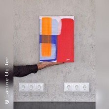 Eine abstrakte Leinwandmalerei mit geometrischen Formen in Rot, Blau und Orange wird von einer Hand vor einer grauen Wand gehalten.