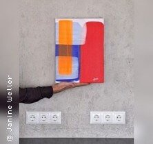 Eine abstrakte Leinwandmalerei mit geometrischen Formen in Rot, Blau und Orange wird von einer Hand vor einer grauen Wand gehalten.