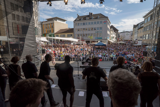 Blick von der Bühne auf den gut besuchten Kiliansplatz. Im Vordergrund ein Chor während des Auftritts.