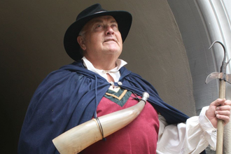 Brustbild des Nachtwächters in Tordurchgang stehend - in rotem Wams, dunkelblauem Unhang und schwarzem Hut, mit Hellebarde in der linken Hand und umgehängtem Horn | © Petra Natzkowski