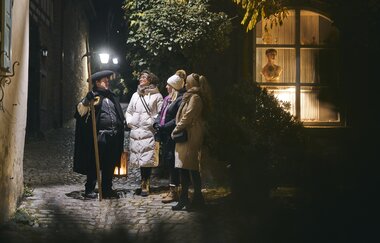 Ein Gruppe Menschen hört gespannt einem Erzähler zu. Es ist Nacht. Der Gästeführer ist als Nachtwächter verkleidet, trägt eine Hellbarde und eine Laterne. | © Tourimia Tourismus GmbH