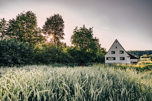 Ein gemütliches Häuschen am Rande eines goldenen Getreidefeldes. Im Hintergrund ragen Bäume empor, während die untergehende Sonne den Himmel in warme Farben taucht. | © Touristikgemeinschaft Odenwald e.V.