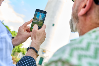 Zwei Herren mit einer Handykamera im Vordergrund, die gerade dabei sind ein Foto vom Hockenheimer Wasserturm zu machen. Auf der Handykamera ist das Gesamtbild des Wasserturms zu sehen. Auf dem Foto erscheint der Wasserturm im Hintergrund. | © Tourimia Tourismus GmbH