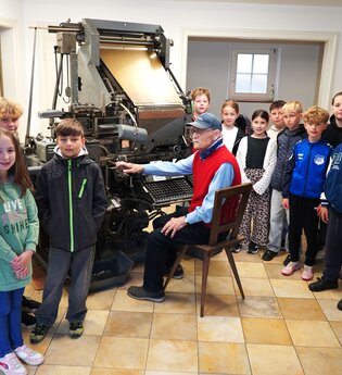 An der Linotype Setzmaschine sitz ein Mann, um ihn herum steht eine Gruppe Kinder. | © Oskar Fritzmann