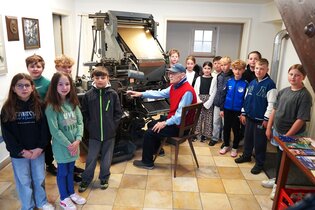 An der Linotype Setzmaschine sitz ein Mann, um ihn herum steht eine Gruppe Kinder. | © Oskar Fritzmann