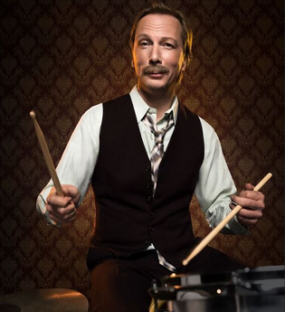 Ein Mann in Weste und Hemd spielt Schlagzeug vor einer gemusterten Tapete. | © C² CONCERTS GmbH