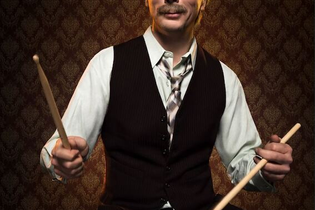 Ein Mann in Weste und Hemd spielt Schlagzeug vor einer gemusterten Tapete. | © C² CONCERTS GmbH