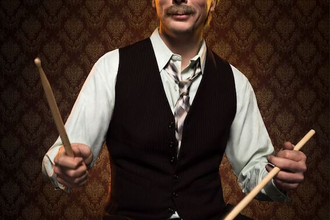 Ein Mann in Weste und Hemd spielt Schlagzeug vor einer gemusterten Tapete. | © C² CONCERTS GmbH