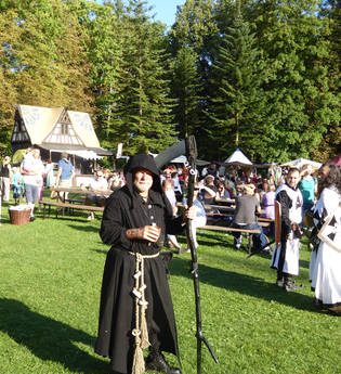 Bärtiger Mann in Mönchskittel mit Sense vor mittelalterlicher Fachwerkhütte mit Holztischen und -bänken | © Landratsamt Rhein-Neckar