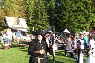 Bärtiger Mann in Mönchskittel mit Sense vor mittelalterlicher Fachwerkhütte mit Holztischen und -bänken | © Landratsamt Rhein-Neckar