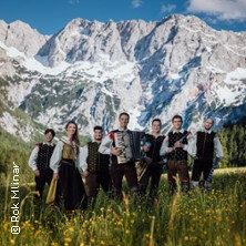 Eine Musikgruppe in traditioneller Kleidung steht auf einer Wiese vor einer Bergkulisse. | © links im Bild
