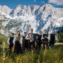 Eine Musikgruppe in traditioneller Kleidung steht auf einer Wiese vor einer Bergkulisse. | © links im Bild