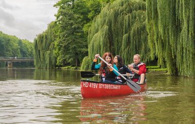 ein rotes Kanu auf dem Neckar. es sitzen 2 Frauen und ein Mann im Boot . Im Hintergrund sieht man eine Brücke. Am Ufersaum stehen hohe Bäume und Trauerweiden