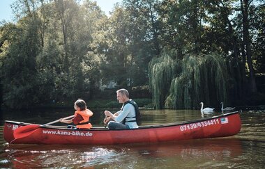 ein Mann und ein Kind fahren in einem roten Kanu auf dem Neckar. Das Kind trägt eine orangene Schwimmweste. Das Kanu wird von zwei Schwänen begleitet. Am Ufersaum stehen hohe Bäume