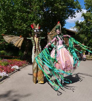 Zwei Personen auf Stelzen in fantasievollen Kostümen in einem blühenden Garten. | © Erlebnispark Tripsdrill