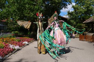 Zwei Personen auf Stelzen in fantasievollen Kostümen in einem blühenden Garten. | © Erlebnispark Tripsdrill
