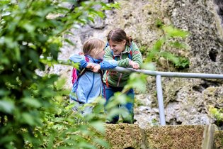 Zwei Kinder lachen gemeinsam vor einer Felswand. Sie tragen Wanderklamotten und haben jeweils einen Wanderrucksack auf. | © Touristikgemeinschaft Hohenlohe e. V. | Florian Trykowski