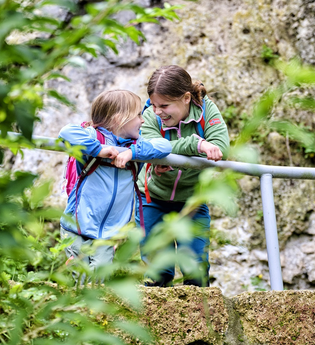 Zwei Kinder lachen gemeinsam vor einer Felswand. Sie tragen Wanderklamotten und haben jeweils einen Wanderrucksack auf. | © Touristikgemeinschaft Hohenlohe e. V. | Florian Trykowski
