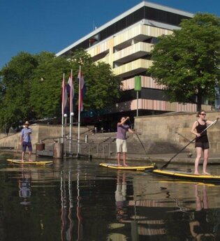 drei Personen auf gelben Paddle Boards. Im Hintergrund die Kaimauer und das Marahaus, teilweise verdeckt durch große Bäume mit grünem Laub. Der Himmel ist blau. Die Personen auf dem Board sind mit Shorts und T-Shirts bekleidet