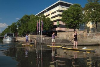 drei Personen auf gelben Paddle Boards. Im Hintergrund die Kaimauer und das Marahaus, teilweise verdeckt durch große Bäume mit grünem Laub. Der Himmel ist blau. Die Personen auf dem Board sind mit Shorts und T-Shirts bekleidet