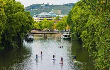 Mehrere Stand-up-Paddler sind in der Mitte des Neckars erkennbar. Die Laubbäume am Neckarsaum sind grün belaubt. In der Ferne ist der Wartberg zu sehen