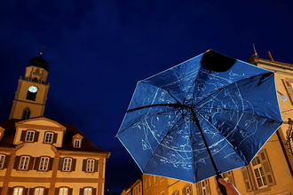 Ein blick in den Nachthimmel. eine Regenschirm wird hochgehalten, indem man die Sternbilder auf der Innenseite sehen kann. Der Kirchturm ist im Hintergrund. | © Stadt Bad Mergentheim