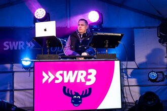 DJ auf Bühne mit Scheinwerfern an Pult mit SWR3 Logo in Pink