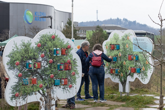 2 Besucher vor dem Gebäude der Klima Arena. Im Vordergrund zwei Plakate in BaumformPlakaten