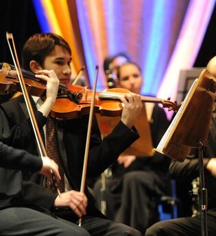 Ein Orchester spielt auf einer Bühne, mehrere Musiker mit Geigen. | © MCS