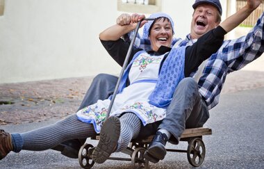 Zwei fröhliche Menschen in traditioneller Kleidung fahren auf einem kleinen Wagen die Straße hinunter. | © Stadt Besigheim