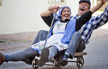 Zwei fröhliche Menschen in traditioneller Kleidung fahren auf einem kleinen Wagen die Straße hinunter. | © Stadt Besigheim