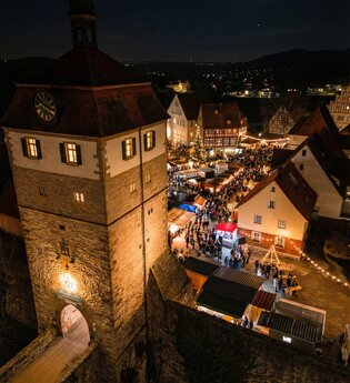 Nachtsicht eines beleuchteten Weihnachtsmarktes mit Menschen, die Stände besuchen, neben einem beleuchteten Turm mit Uhr.