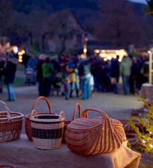 Im Vordergrund sind mehrere Weidenkörbe und auf der rechten Seite ein beleuchteter Nadelbaum. Im Hintergrund sind Menschen und  verschiedene Marktstände.