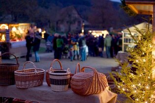 Im Vordergrund sind mehrere Weidenkörbe und auf der rechten Seite ein beleuchteter Nadelbaum. Im Hintergrund sind Menschen und  verschiedene Marktstände.