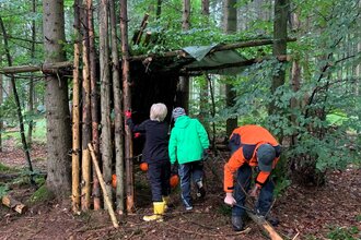 Drei Personen bauen eine Hütte aus Ästen und Zweigen im Wald. | © Forstamt Schwäbisch Hall
