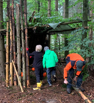 Drei Personen bauen eine Hütte aus Ästen und Zweigen im Wald. | © Forstamt Schwäbisch Hall