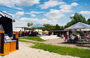 Strandkorb auf Sandfläche, Personen sitzen an Kaffeehaustischen und Biertischen | © Heuchelberg Weingärtner eG