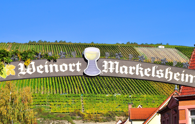 Ein Schild mit der Aufschrift "Weinort Markelsheim", dekoriert mit einem Weinglas. Im Hintergrund sieht man die Weinberge. | © Thomas Weller