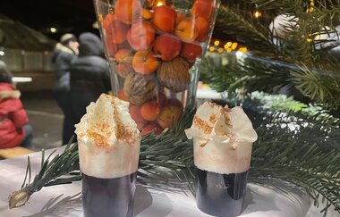 Zwei heiße Getränke mit Sahne und Zimt, dahinter eine Vase gefüllt mit Nüssen und kleinen Äpfeln. Im Hintergrund befinden sich Tannenzweige und Lichter als Dekoration.