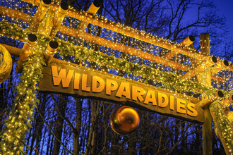 Eingangstor mit der Aufschrift 'Wildparadies', geschmückt mit Lichterketten und Tannenzweigen, vor einem dunklen Himmel. | © Erlebnispark Tripsdrill