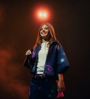Die Künstlerin Zah1de steht auf der Bühne und hält ein Mikrofon in der Hand. Im Hintergrund leuchtet ein Scheinwerfer. | © William Krause