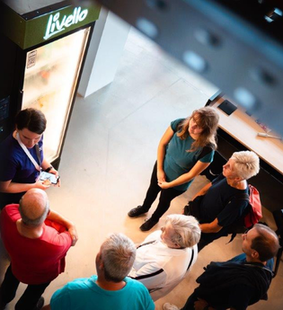 Das Bild zeigt eine Gruppe Menschen aus der Vogelperspektive, die sich um einen Kühlschrank-Automaten versammelt haben. Eine Person aus dem Halbkreis erklärt etwas dazu. Die Gruppe wirkt interessiert.