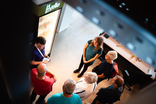 Das Bild zeigt eine Gruppe Menschen aus der Vogelperspektive, die sich um einen Kühlschrank-Automaten versammelt haben. Eine Person aus dem Halbkreis erklärt etwas dazu. Die Gruppe wirkt interessiert.