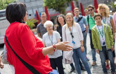 Eine Frau in roter Bluse, die mit dem Rücken zur Kamera steht, mit Headset-Mikrofont, spricht und gestikuliert in Richtung einer Gruppe. In der Gruppe trägt jeder eine Art Schlüsselband mit einem Pass um den Hals. Die Teilnehmer hören aufmerksam zu.