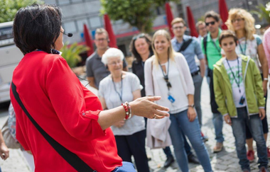 Eine Frau in roter Bluse, die mit dem Rücken zur Kamera steht, mit Headset-Mikrofont, spricht und gestikuliert in Richtung einer Gruppe. In der Gruppe trägt jeder eine Art Schlüsselband mit einem Pass um den Hals. Die Teilnehmer hören aufmerksam zu.